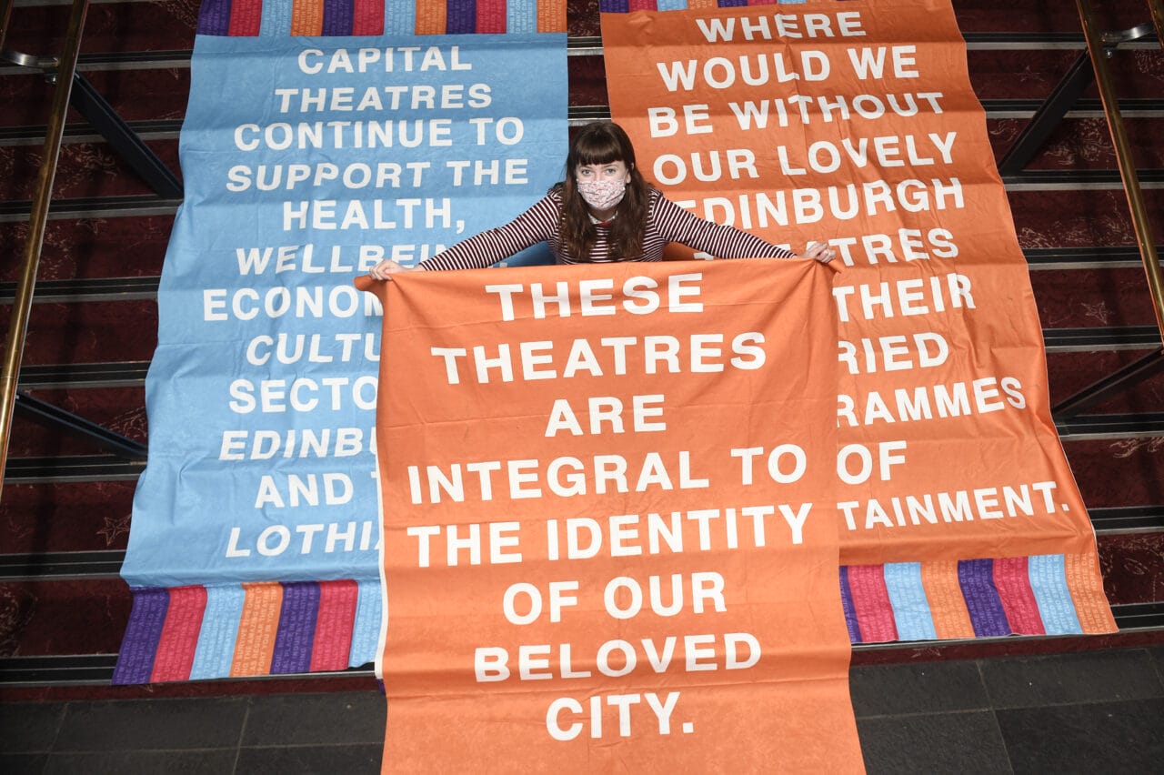 A person holds a large orange banner on stairs, surrounded by other banners with messages supporting the importance of theatres to Edinburgh's identity and well-being.