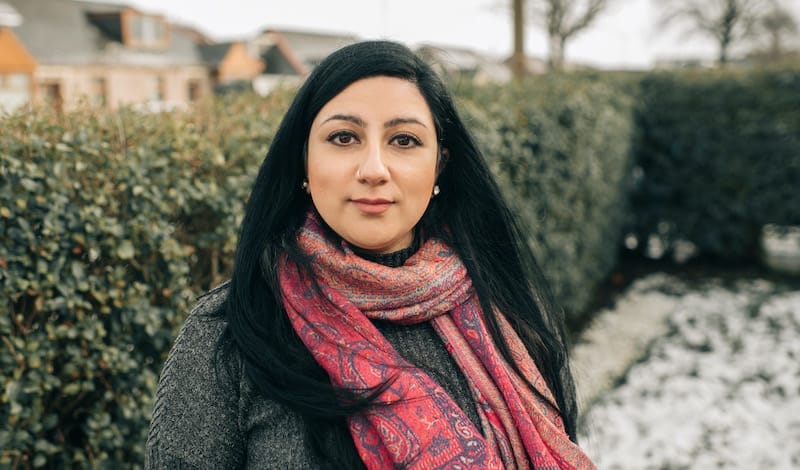 Niloo Far-Khan, a woman with long dark hair wearing a patterned pink scarf and gray coat stands outside near hedges and snow-covered ground.