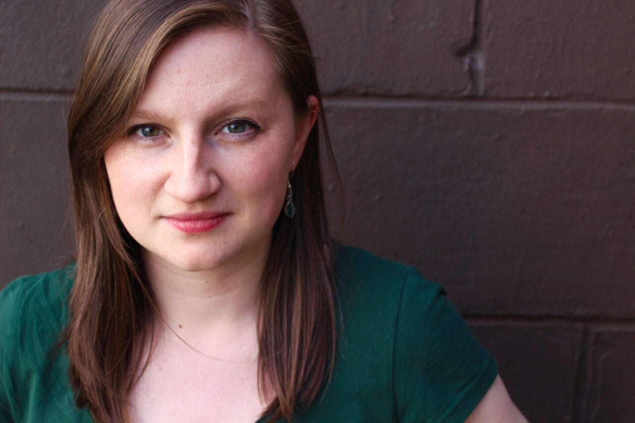 Shonagh Murray, a woman with straight brown hair and a green shirt, looks at the camera while standing in front of a brown brick wall.