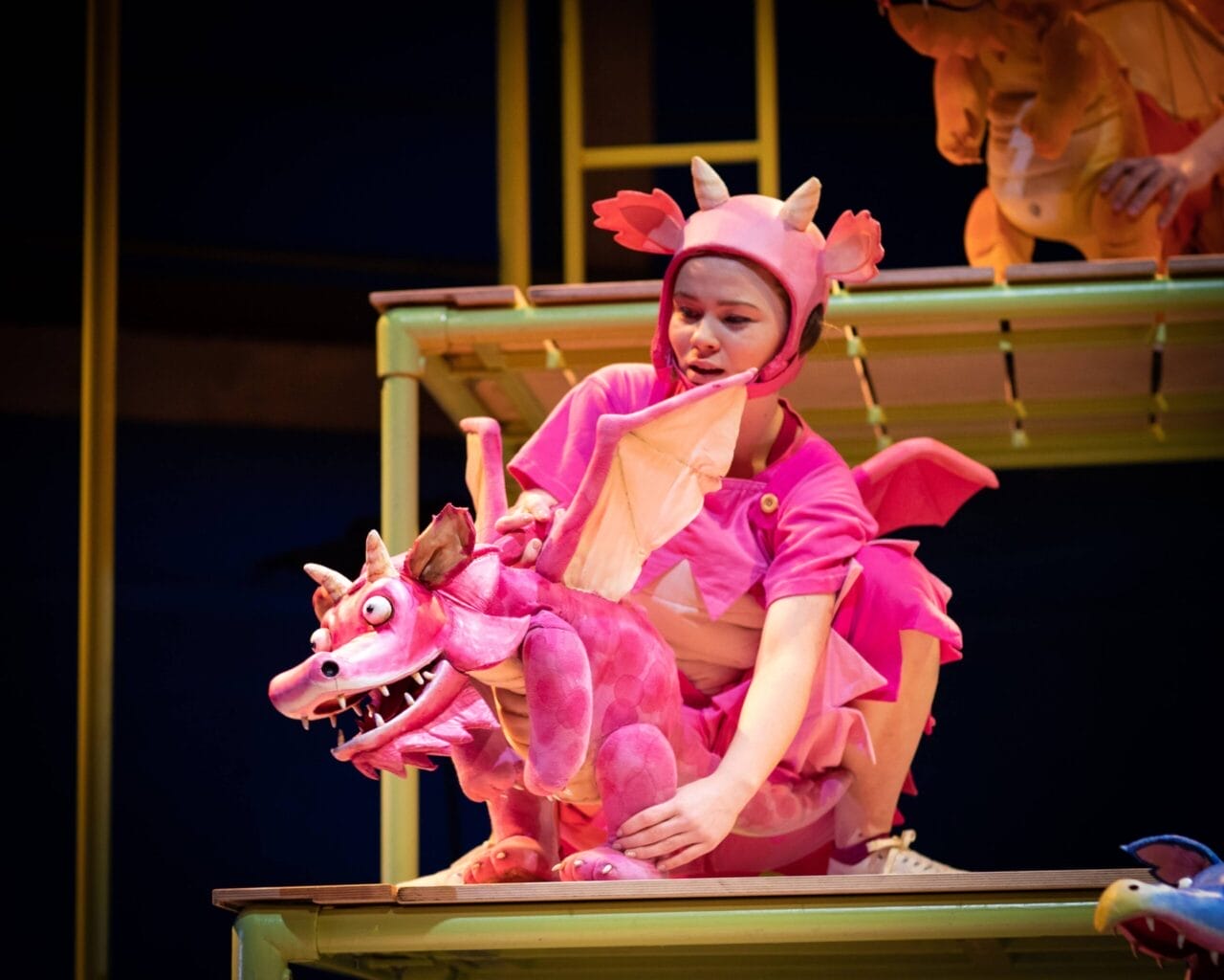 A person in a pink dragon costume operates a pink dragon puppet under the spotlight on a stage set with yellow-green railings.