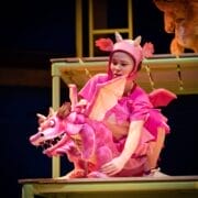 A person in a pink dragon costume operates a pink dragon puppet under the spotlight on a stage set with yellow-green railings.