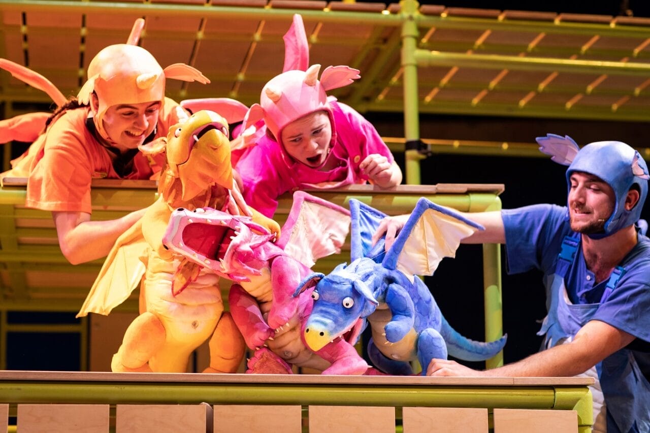 Three people dressed in colorful costumes use dragon puppets—orange, pink, and blue—on a stage with green railings and a yellow backdrop.