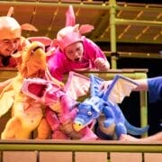Three people dressed in colorful costumes use dragon puppets—orange, pink, and blue—on a stage with green railings and a yellow backdrop.