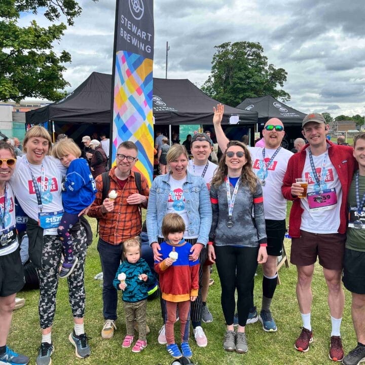 A group of people, some wearing medals and race bibs, pose outdoors at an event with tents and a crowd in the background. Two children stand at the front.