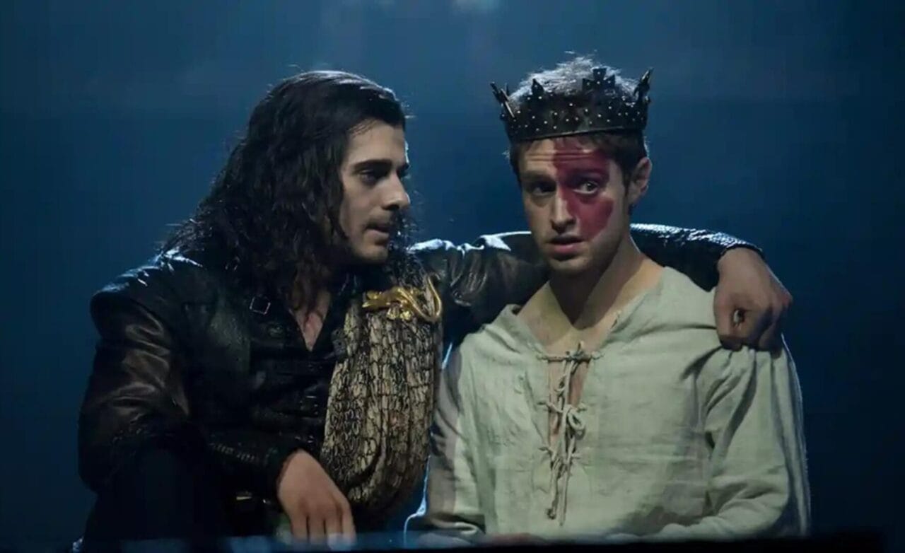 Two men sit together; Daniel Cahill, wearing a crown and white shirt with red paint on his face, is joined by another in darker clothing who has his arm around him and appears to be talking, both captured in the spotlight.
