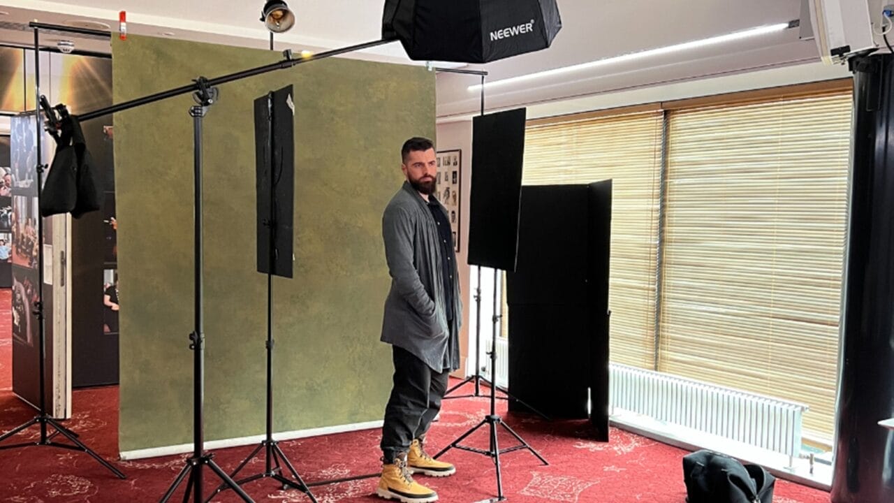 Daniel Cahill stands in a photography studio setup with lighting equipment, a green backdrop, and closed window blinds.