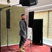 Daniel Cahill stands in a photography studio setup with lighting equipment, a green backdrop, and closed window blinds.