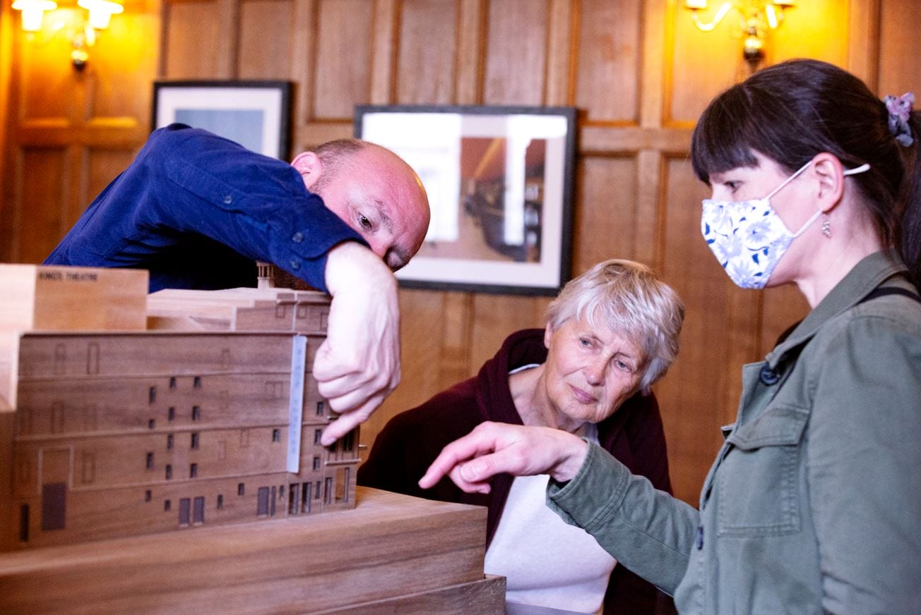 A man gestures to details on a wooden architectural model while an older woman and a younger woman wearing a mask closely observe and point.