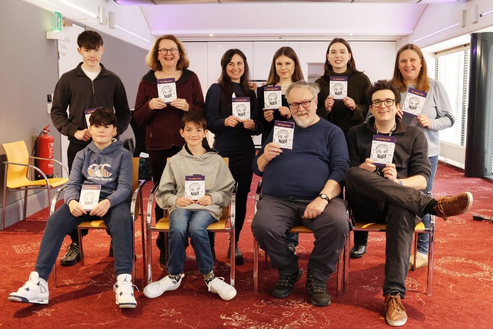 A group of ten people, adults and children, pose indoors holding matching booklets while seated and standing in two rows on a red carpeted floor.