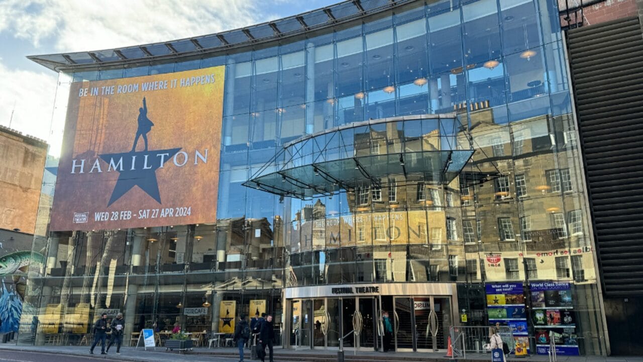 Modern glass-fronted theater building with large "Hamilton" musical poster and reflection of surrounding cityscape, people walking in front.