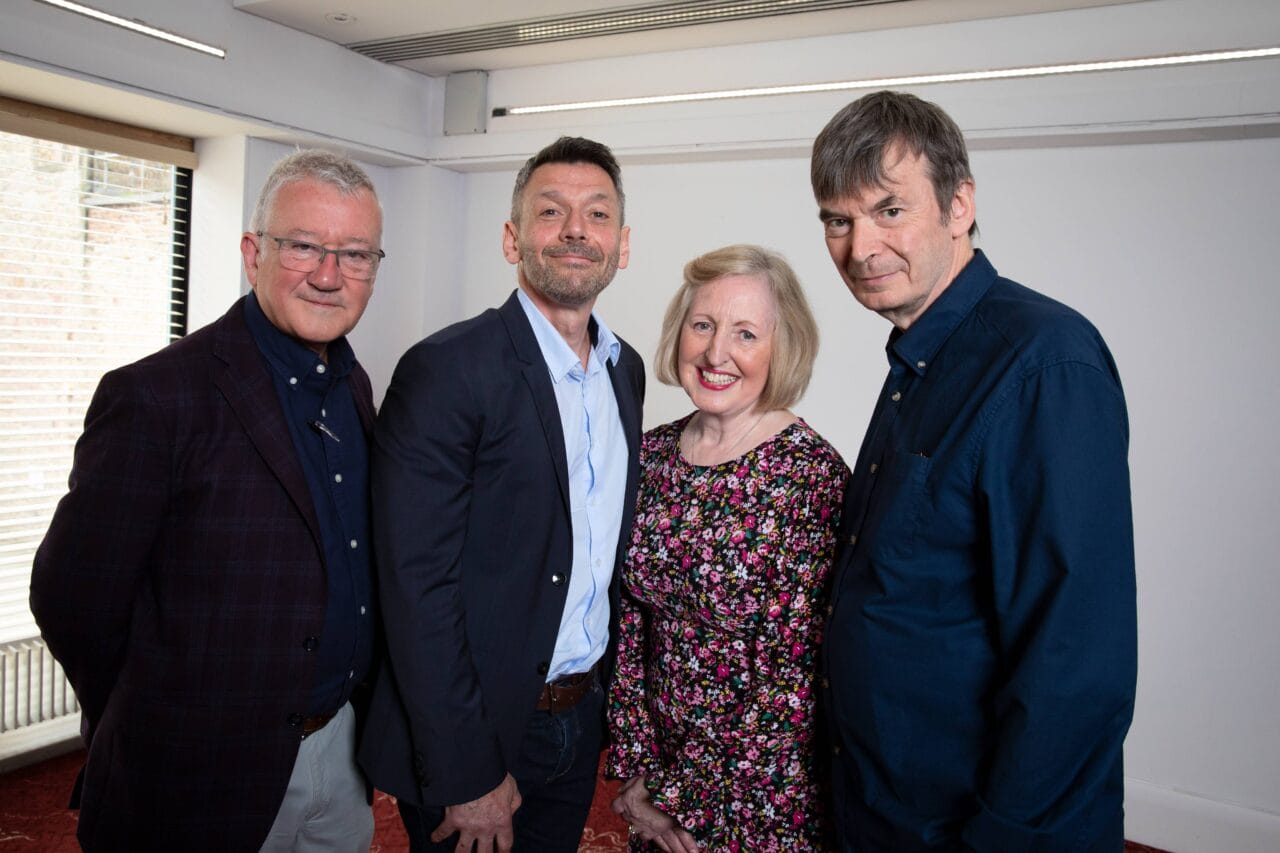 Four adults, three men and one woman, stand together indoors, posing for a group photo. They are dressed in smart-casual clothing and smiling at the camera.