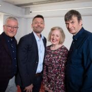 Four adults, three men and one woman, stand together indoors, posing for a group photo. They are dressed in smart-casual clothing and smiling at the camera.