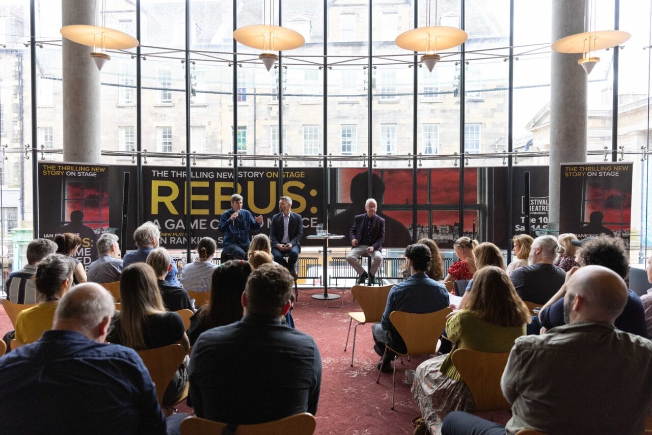 A group of people seated in a room listen to two speakers on a stage with a large "Rebus: The Story on Stage" banner in the background.