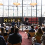 A group of people seated in a room listen to two speakers on a stage with a large "Rebus: The Story on Stage" banner in the background.