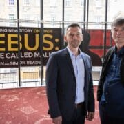 Two men in business-casual attire stand indoors in front of large promotional posters for "REBUS: A Game Called Malice.