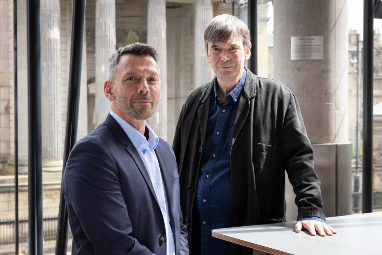 Two middle-aged men stand indoors near a table, with columns and large windows in the background. One man wears a suit, the other wears a dark jacket over a blue shirt.