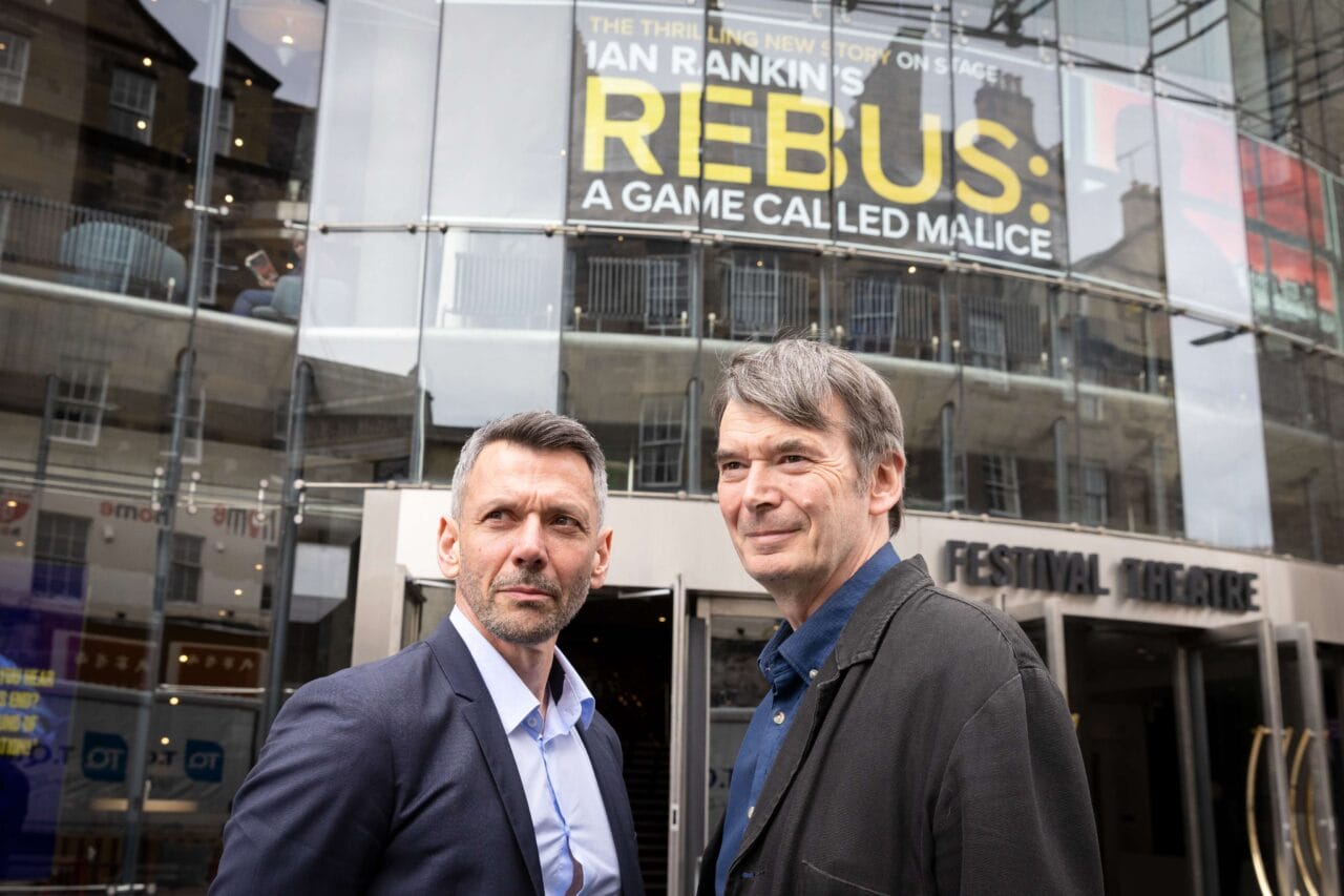 Two men standing outside the Festival Theatre, with a sign above them reading "Rebus: A Game Called Malice" by Ian Rankin.