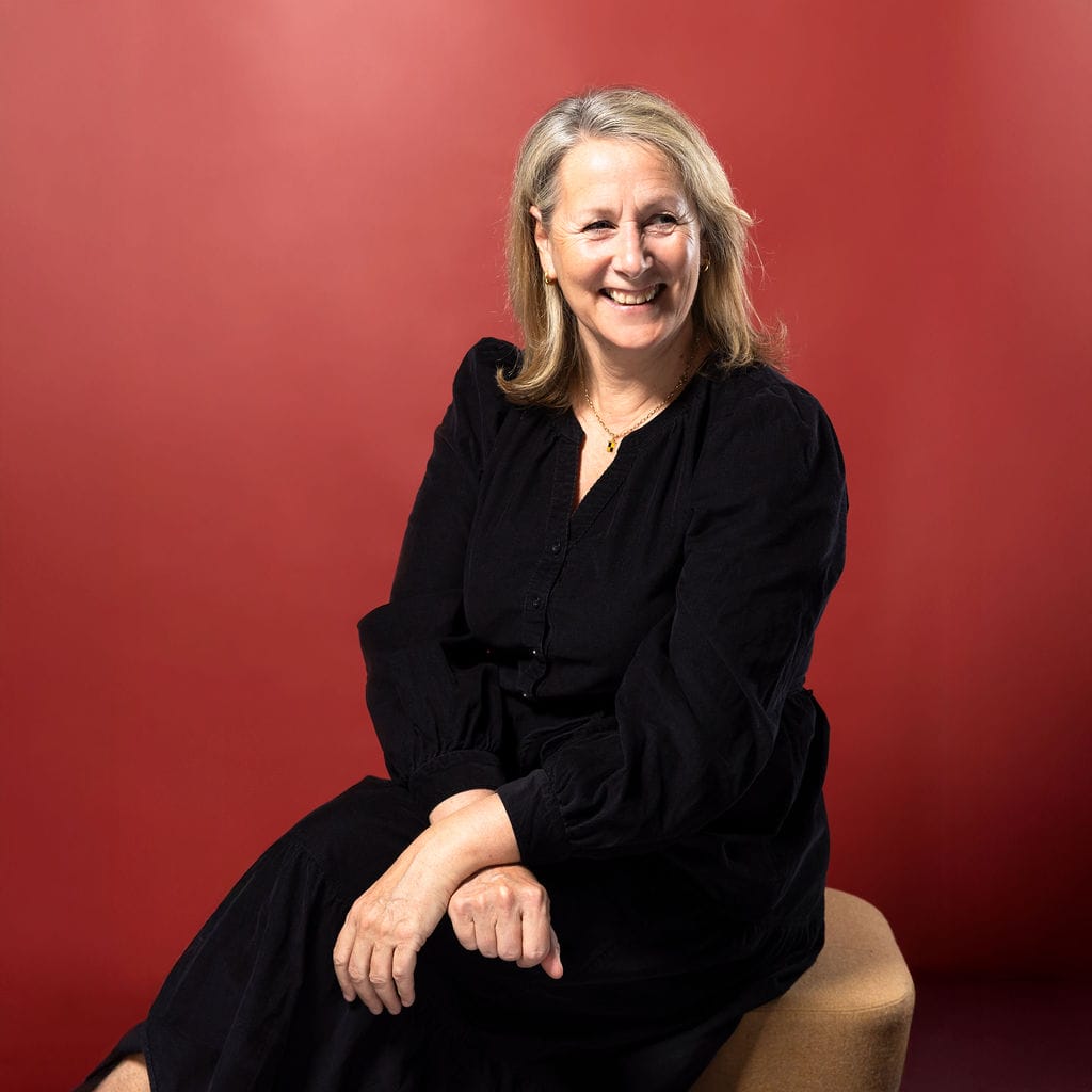 A woman with blonde hair wearing a black dress sits on a stool and smiles, against a plain red background.