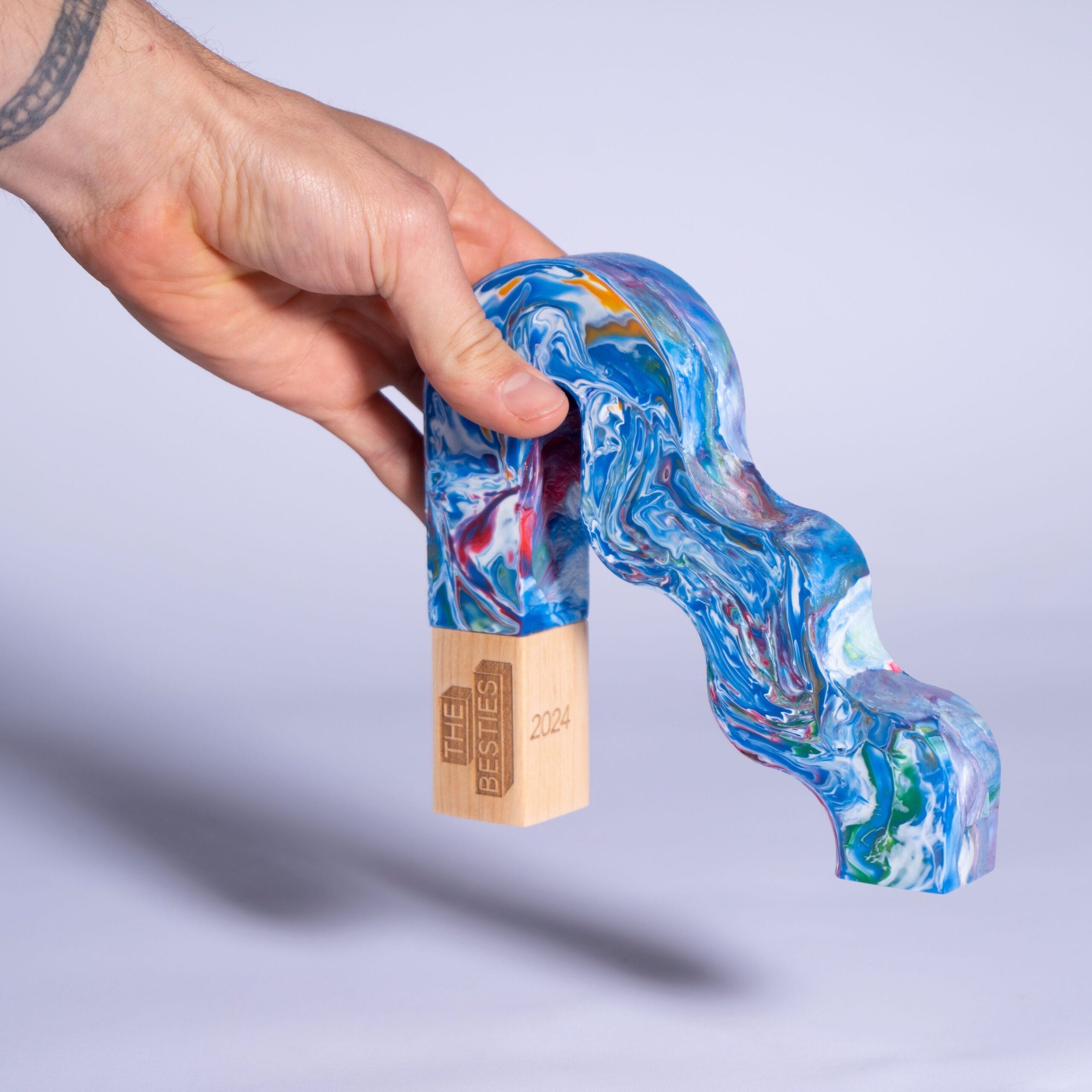 A hand holds a colourful, wavy award made of marbled blue material with a wooden base labelled "THE BESTIES 2024.