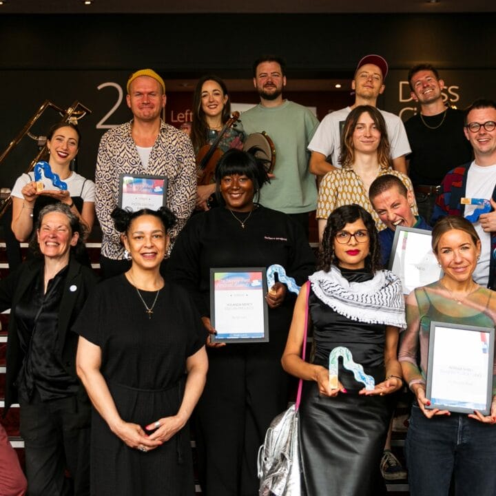 A group of people stand on stairs, holding certificates and blue dinosaur-shaped trophies, posing for a photo at an indoor event.