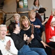 A group of older adults sit together indoors, some holding musical instruments and clapping, while one woman raises her arm enthusiastically; coffee cups are on the tables.