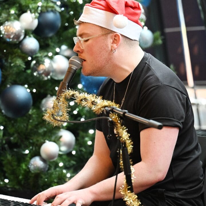 A person wearing a Santa hat plays a keyboard and sings into a microphone near a decorated Christmas tree with blue and silver ornaments.