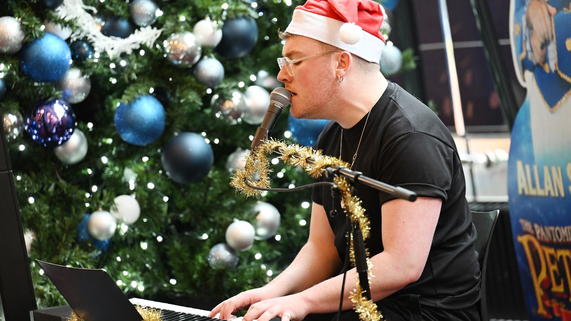 A person wearing a Santa hat plays a keyboard and sings into a microphone near a decorated Christmas tree with blue and silver ornaments.