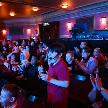 Children and adults sit in theatre seats, some clapping and smiling, while others watch the stage; staff are seen at a control booth in the background.