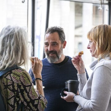 Three adults stand together indoors, eating snacks and drinking coffee while having a conversation. Large windows and urban buildings are visible in the background.
