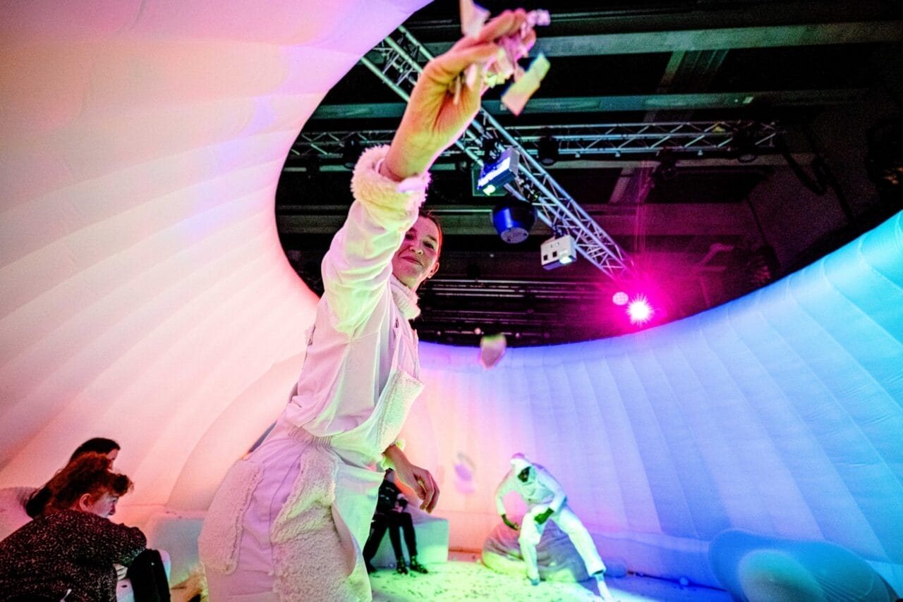 A performer in white brings a sensory production to life, tossing confetti inside an inflatable, color-lit dome as people watch from the side.