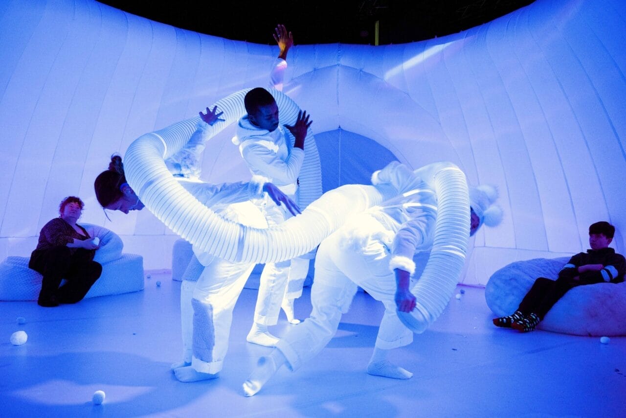 Three performers in white costumes with flexible tubes dance in a blue-lit, futuristic room evoking a sensory production of winter’s chill, while two seated people watch.