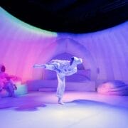 Two people in white costumes are inside a dome-like, inflatable set with soft, colorful lighting; one is sitting and the other is mid-kick in a performance pose during this immersive winter sensory production.