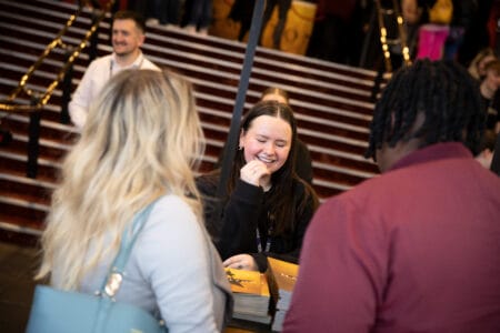 A woman stands behind a table handing out programs to two people at the bottom of a staircase in a busy indoor setting.