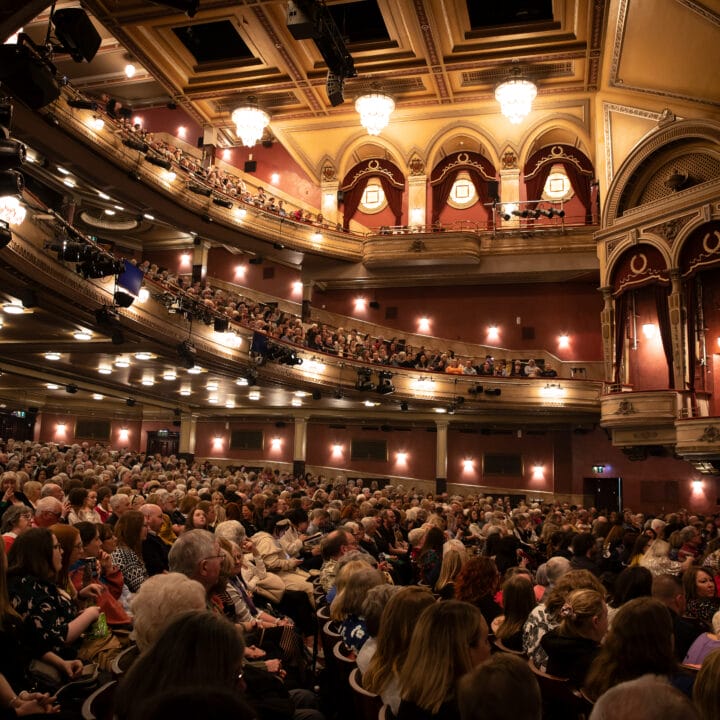 A large audience fills the seats of an ornate theatre with multiple balconies, waiting for a performance to begin.