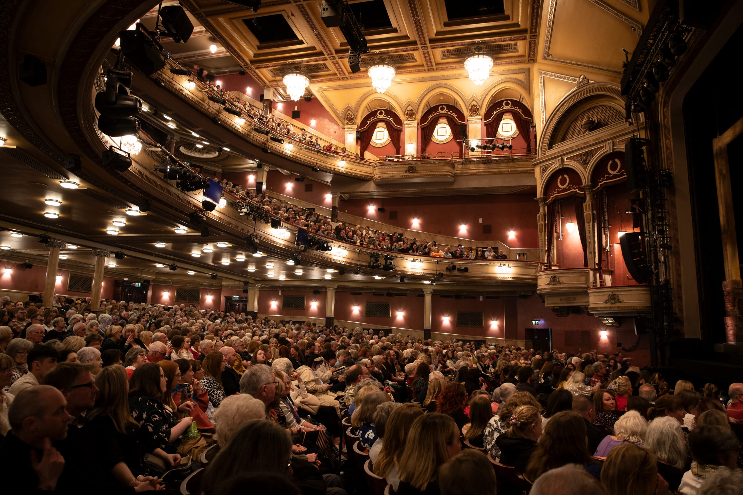 A large audience fills the seats of an ornate theatre with multiple balconies, waiting for a performance to begin.