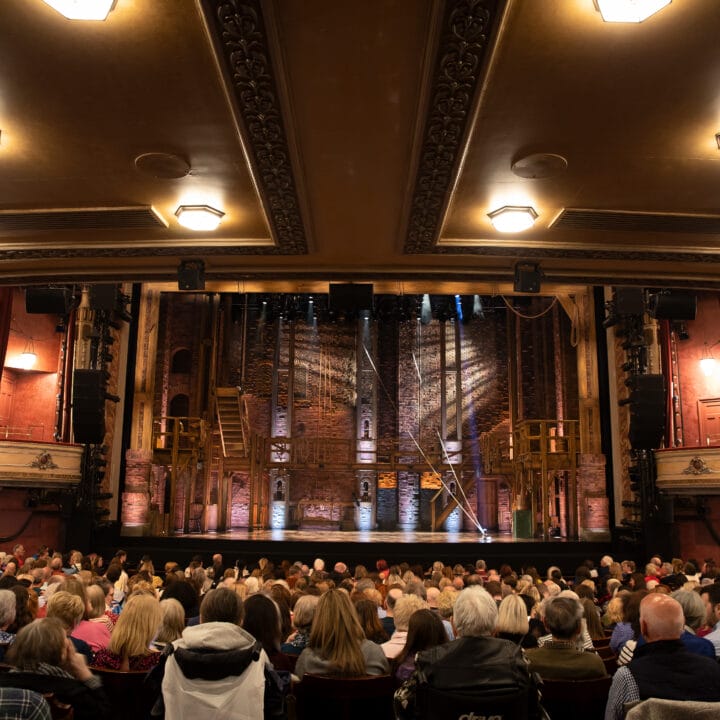 A full audience sits in a historic theatre facing a stage set with wooden scaffolding and brick walls, awaiting the start of a performance.
