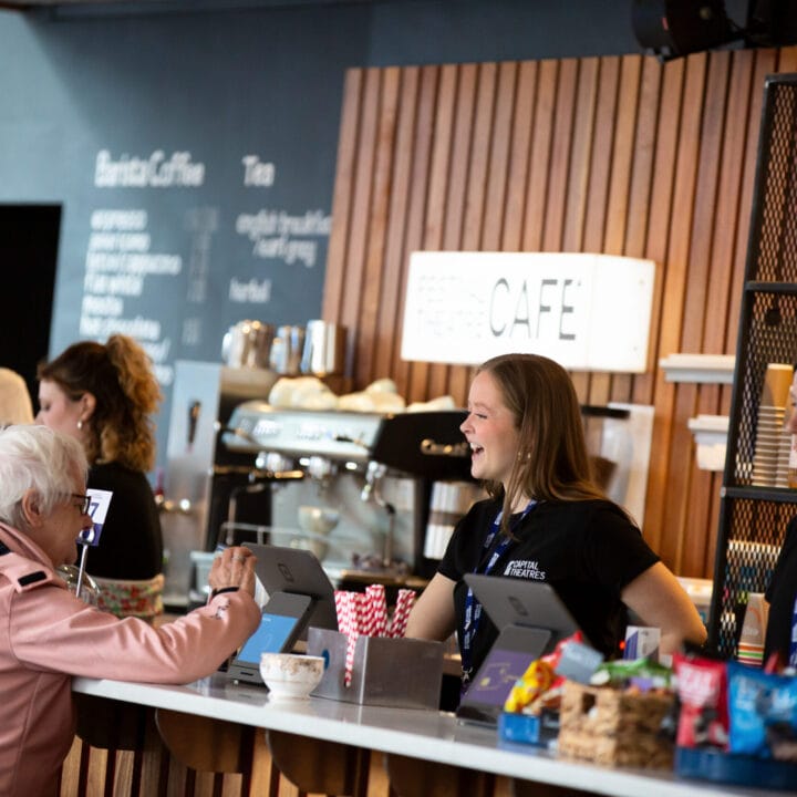 A customer interacts with staff at a modern café counter, where employees serve drinks and snacks, with a coffee machine and menu visible in the background.
