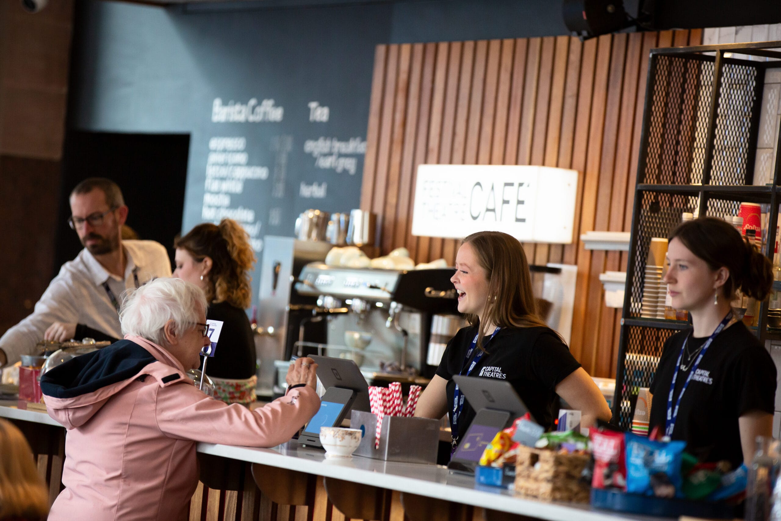 A customer interacts with staff at a modern café counter, where employees serve drinks and snacks, with a coffee machine and menu visible in the background.