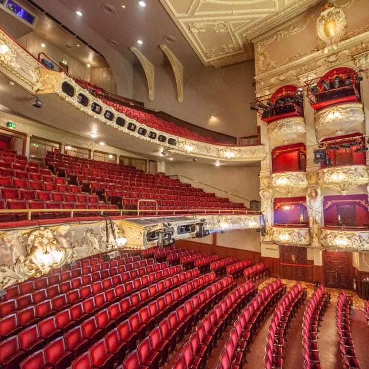 Interior of an ornate theatre with red seats, gold detailing, private box balconies, and a view of the stage area from the audience perspective.