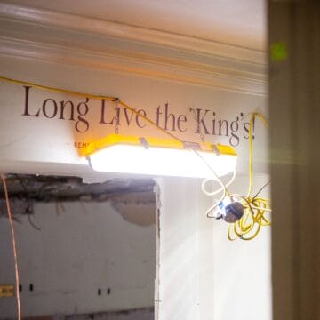 A construction light with exposed wires hangs from a ceiling near a wall with the phrase “Long Live the King’s” in decorative lettering.