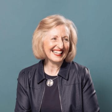Exec Fiona Gibson, a woman with short blonde hair, wears a dark jacket and a round pendant necklace, smiling and looking to the side against a plain blue background.