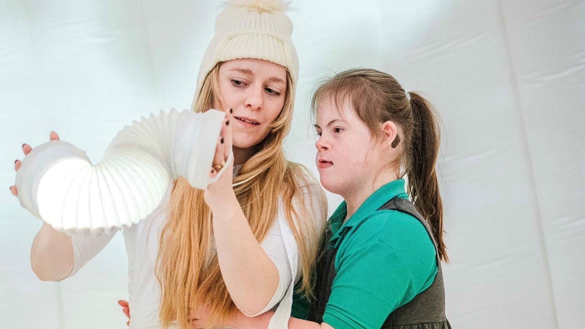 Two girls stand together examining a flexible white light fixture in a bright, white room.