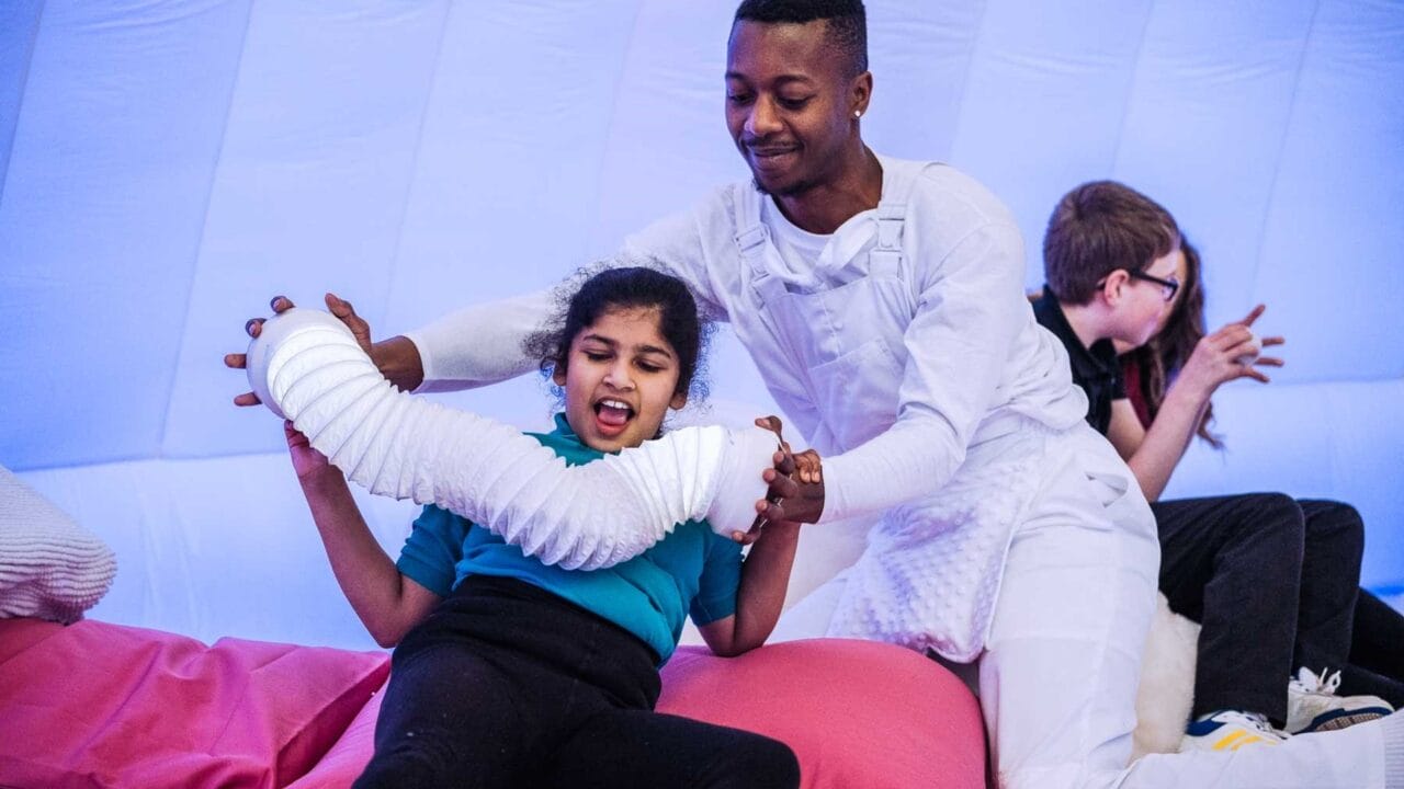 A man in white helps a young girl interact with a flexible white tube in a brightly lit, padded room, while another child and adult are in the background.