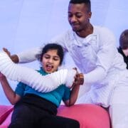 A man in white helps a young girl interact with a flexible white tube in a brightly lit, padded room, while another child and adult are in the background.