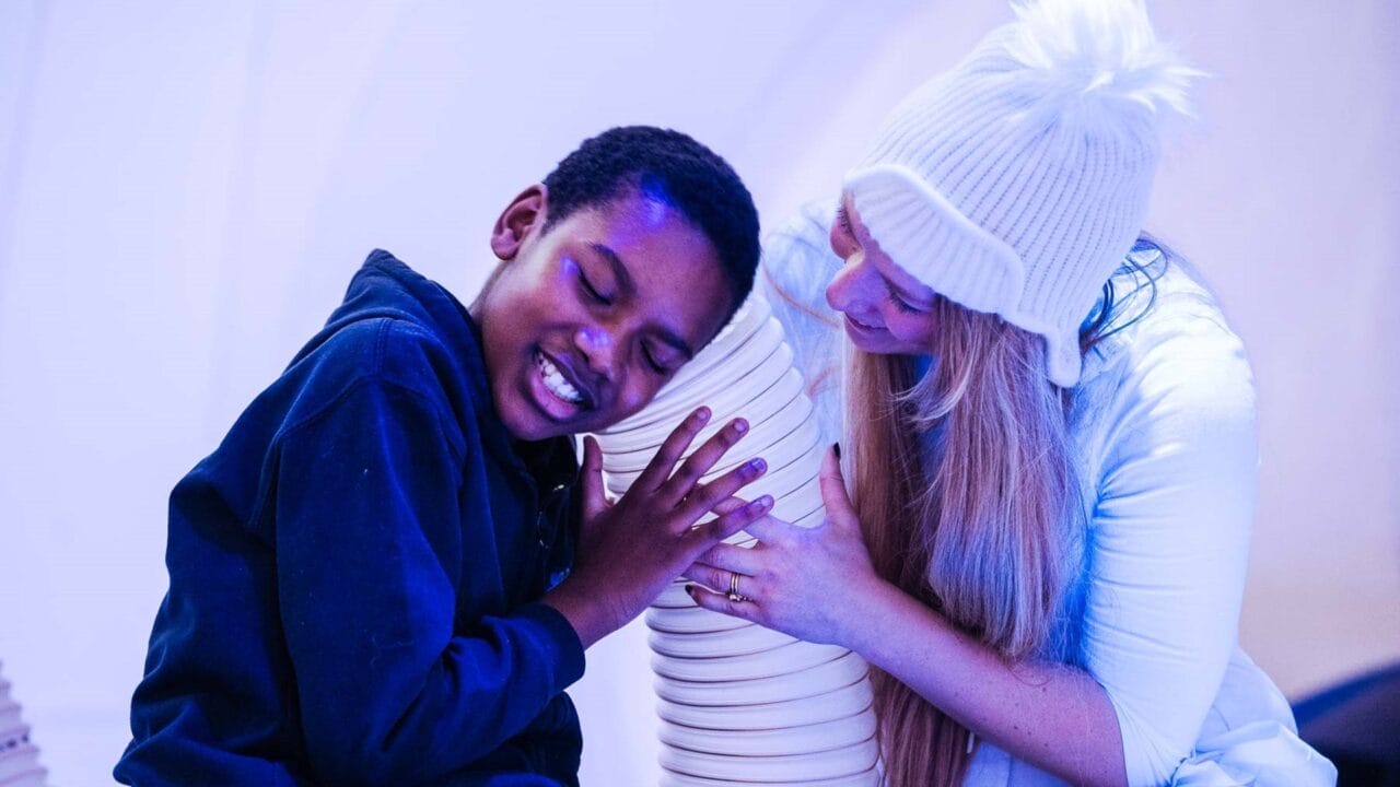 A boy smiles with eyes closed, hugging a large white tube, while a woman in a white hat and sweater kneels beside him, smiling.