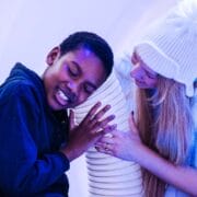 A boy smiles with eyes closed, hugging a large white tube, while a woman in a white hat and sweater kneels beside him, smiling.