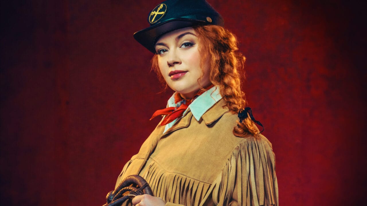 A woman dressed in a fringed tan jacket and black hat with a yellow insignia holds a coiled rope against a red background.
