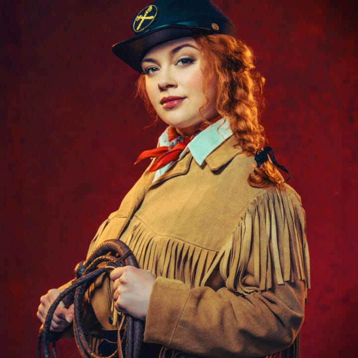 A woman dressed in a fringed tan jacket and black hat with a yellow insignia holds a coiled rope against a red background.