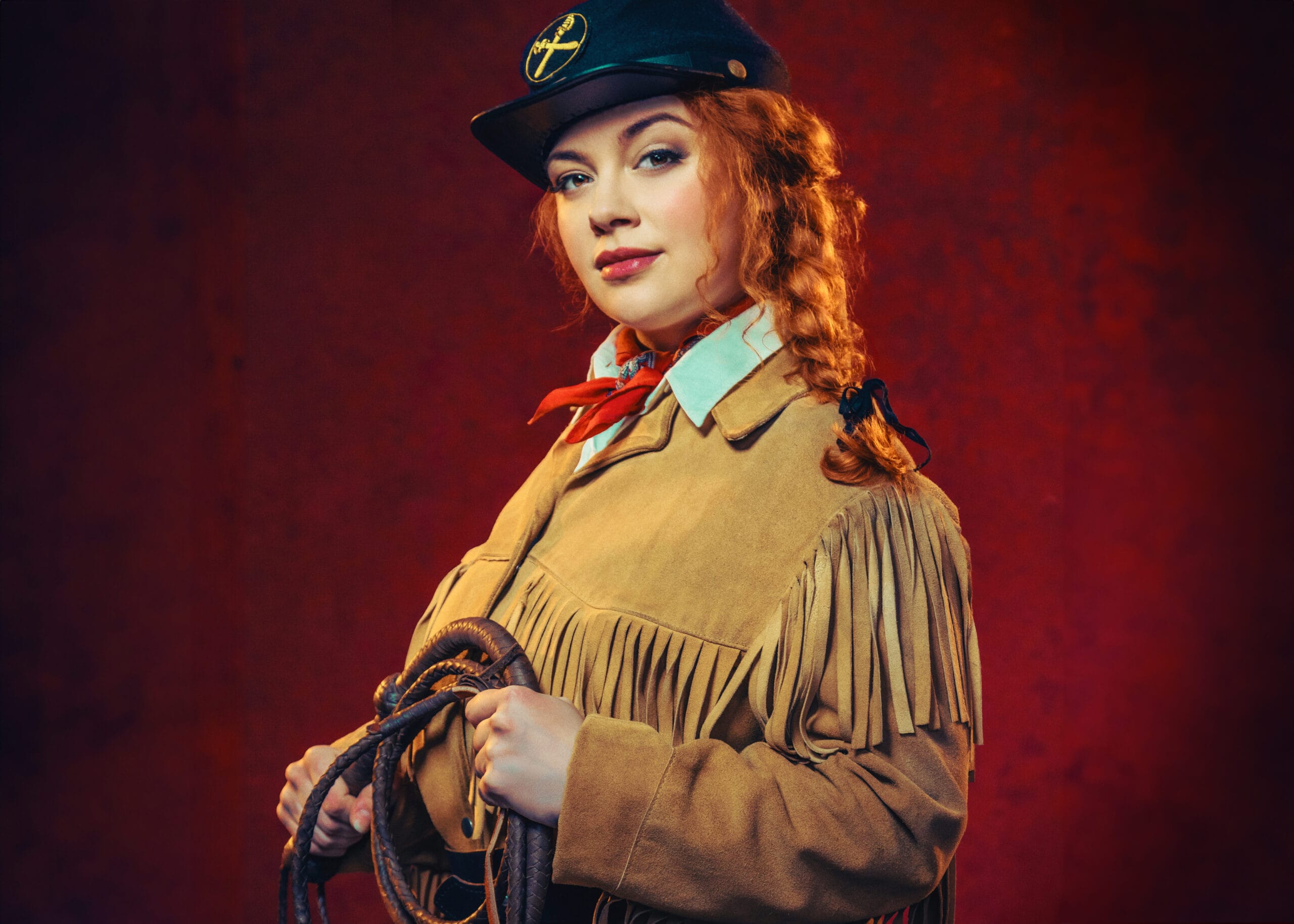 A woman dressed in a fringed tan jacket and black hat with a yellow insignia holds a coiled rope against a red background.