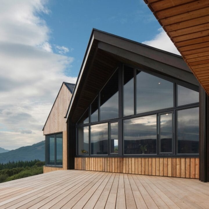 Modern wooden house with large glass windows and a spacious deck, overlooking a scenic landscape with mountains and water under a partly cloudy sky.